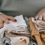 Organized Living - Crop faceless female with manicure arranging baby clothes on warm scarf while sitting at table in light room at home