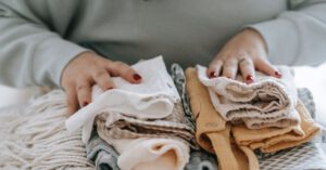 Organized Living - Crop faceless female with manicure arranging baby clothes on warm scarf while sitting at table in light room at home