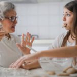 Elderly Home - Calm senior woman and teenage girl in casual clothes looking at each other and talking while eating cookies and cooking pastry in contemporary kitchen at home