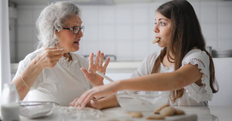 Elderly Home - Calm senior woman and teenage girl in casual clothes looking at each other and talking while eating cookies and cooking pastry in contemporary kitchen at home