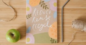 Reuse Art - Top view of book with Reduce Reuse Recycle inscriptions near green apple and bobbin of thread on table