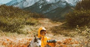 Pets Weather - Full body frozen traveler resting on grassy highland with adorable Husky dog and pouring hot drink from thermos bottle against majestic snowy mountain range on cold day