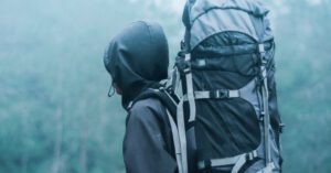 Travel Safety - Man Wearing Black Hoodie Carries Black and Gray Backpacker Near Trees during Foggy Weather