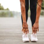 Travel Health - From below crop slender female athlete in sportswear and white sneakers doing standing forward bend exercise for stretching body on wooden floor of street sports ground against blurred urban environment in daytime