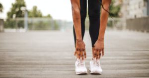 Travel Health - From below crop slender female athlete in sportswear and white sneakers doing standing forward bend exercise for stretching body on wooden floor of street sports ground against blurred urban environment in daytime