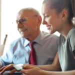 Smart Finance - Young woman in casual clothes helping senior man in formal shirt with paying credit card in Internet using laptop while sitting at table