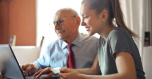 Smart Finance - Young woman in casual clothes helping senior man in formal shirt with paying credit card in Internet using laptop while sitting at table