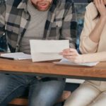 Couples Finance - A Couple Sitting Near the Wooden Table while Looking at the Document in Shocked Emotion